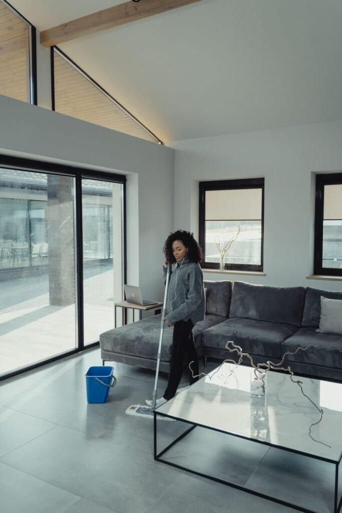 A woman cleans a sleek modern living room with large windows and a mop.